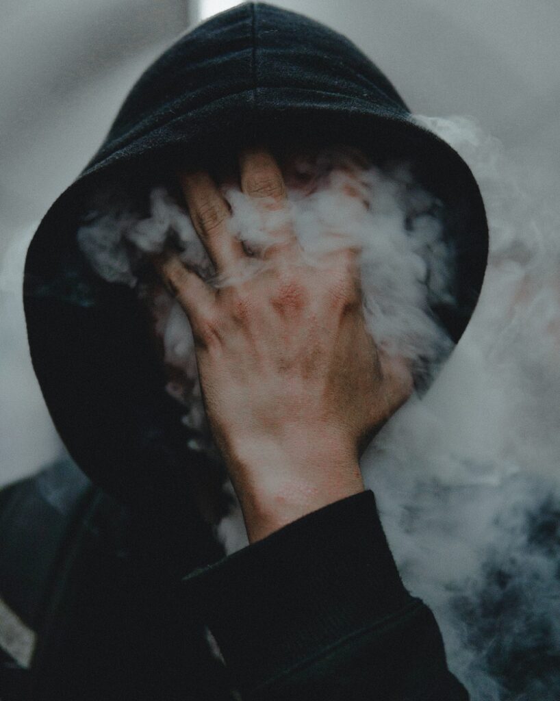 A hooded man with smoke pouring out from his hood. His hand is covering his face. He may be disappearing, it's rather magical.