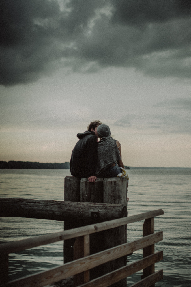 A couple kissing romantically on an ocean dock with gray clouds above.