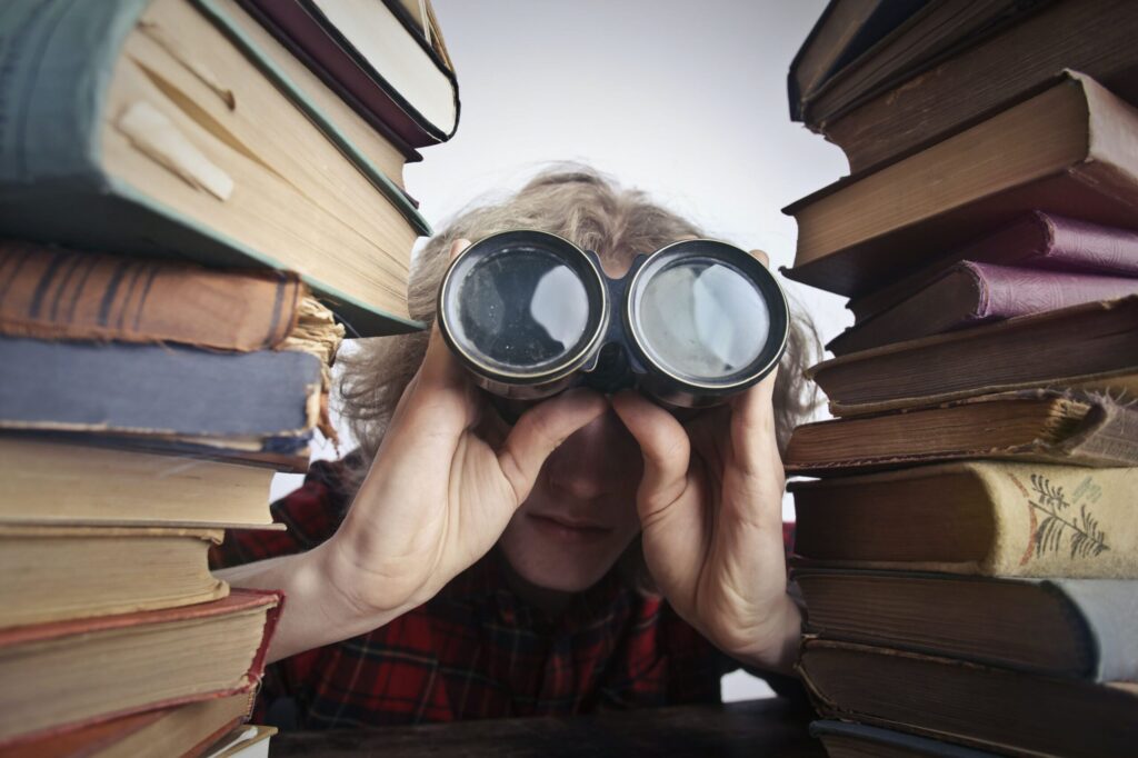 person between stacks of books looking through binoculars for character inspiration